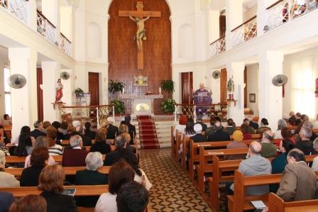 zoomed out picture of the inside of the church, with the cross at the centre visible and church goers praying idoly 